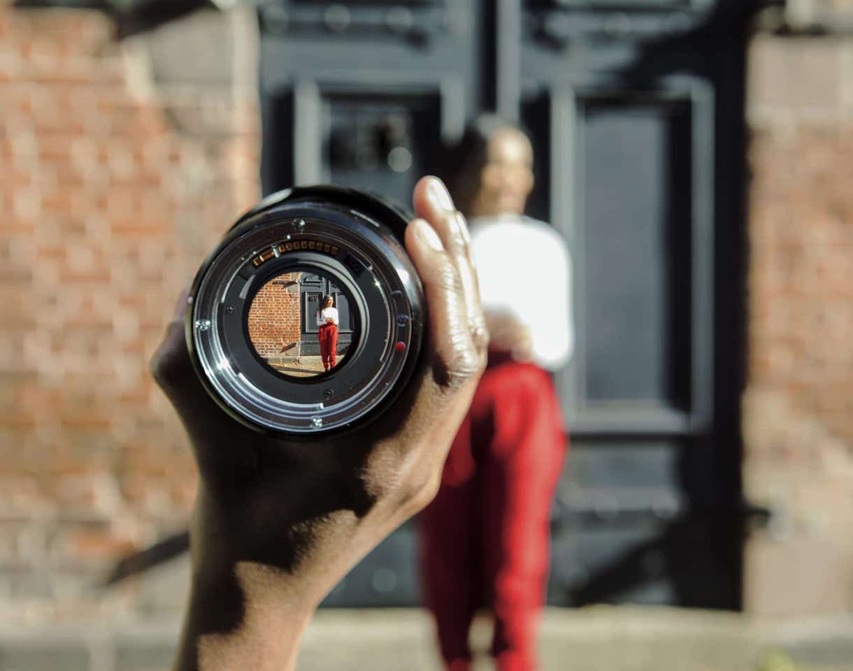 Person out of focus, with smaller image visible through camera lens held in hand