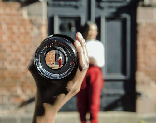 Person out of focus, with smaller image visible through camera lens held in hand